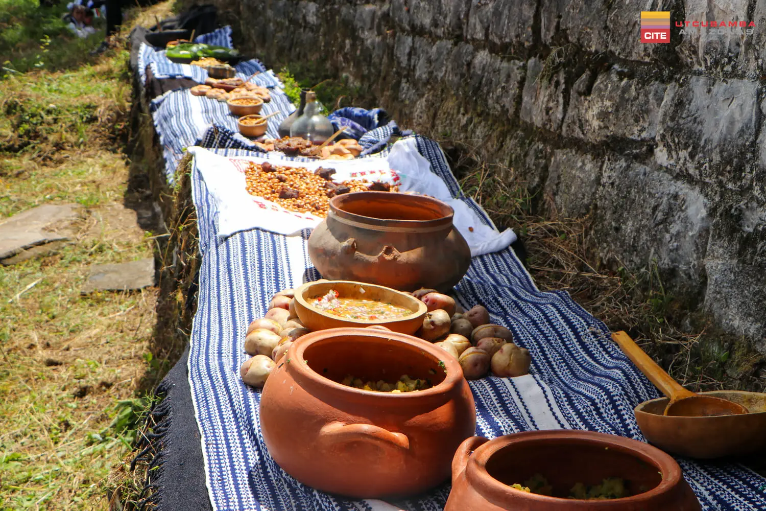 Ollas de barro, papas y comidas servidas sobre mantas tejidas junto a un muro de piedra