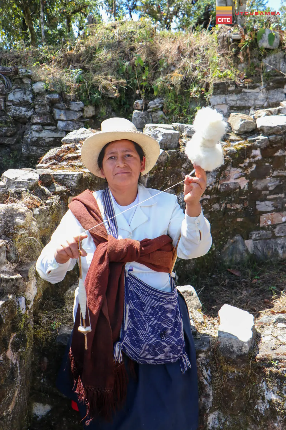 Artesana con sombrero hilando lana con pushcana entre ruinas de piedra