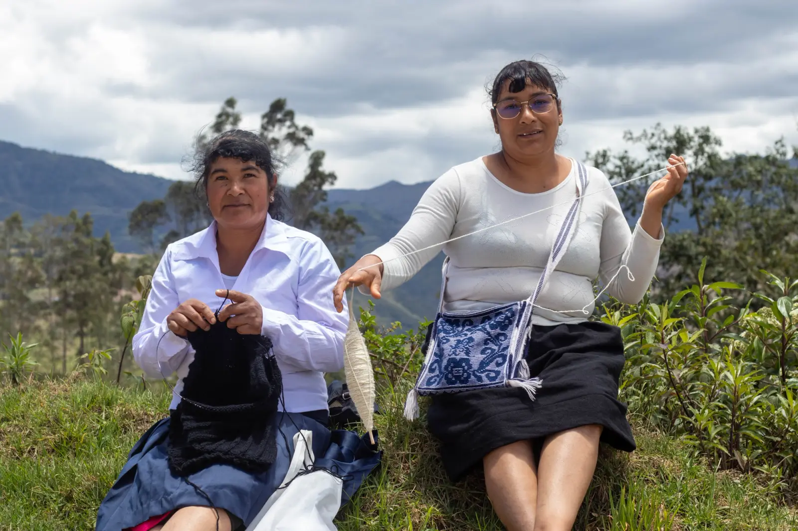 Dos artesanas tejiendo a palillo e hilando lana en una ladera con cerros al fondo
