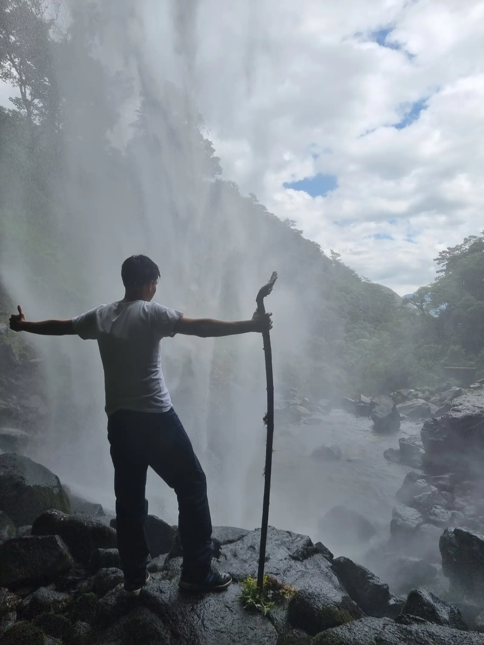 Hombre con bastón de madera sobre rocas, bajo la brisa de la catarata Yumbilla