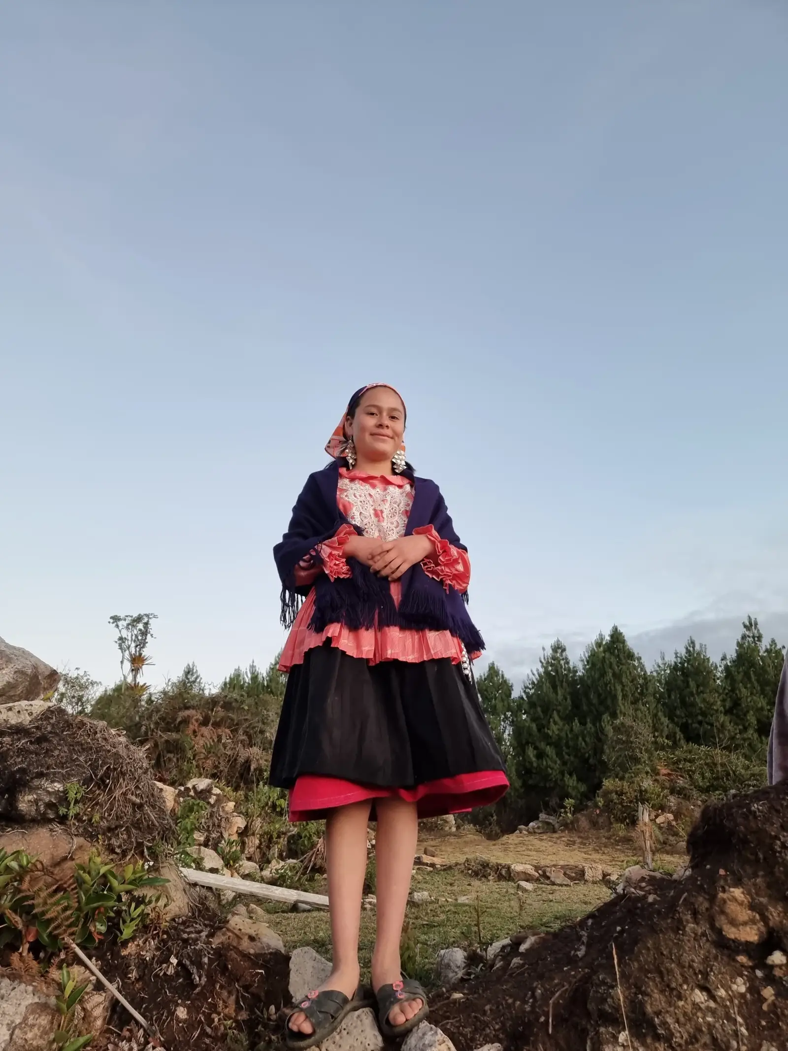 Joven con vestimenta tradicional, blusa bordada y manta azul, junto a un pinar