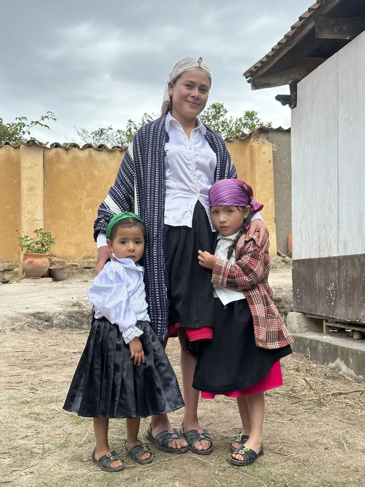Mujer con manta tejida abraza a dos niñas con ropa tradicional en un patio