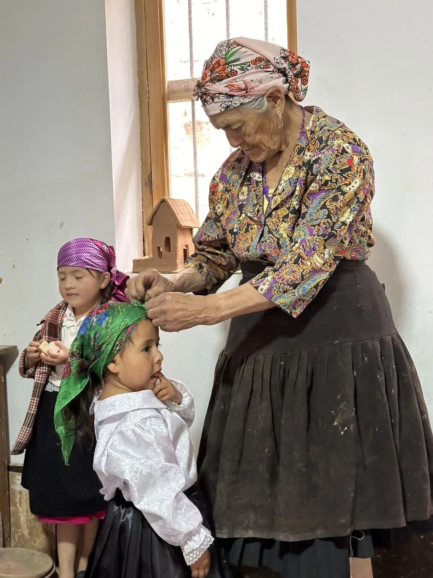 Mujer mayor acomodando el pañuelo de una niña junto a una casita de cerámica