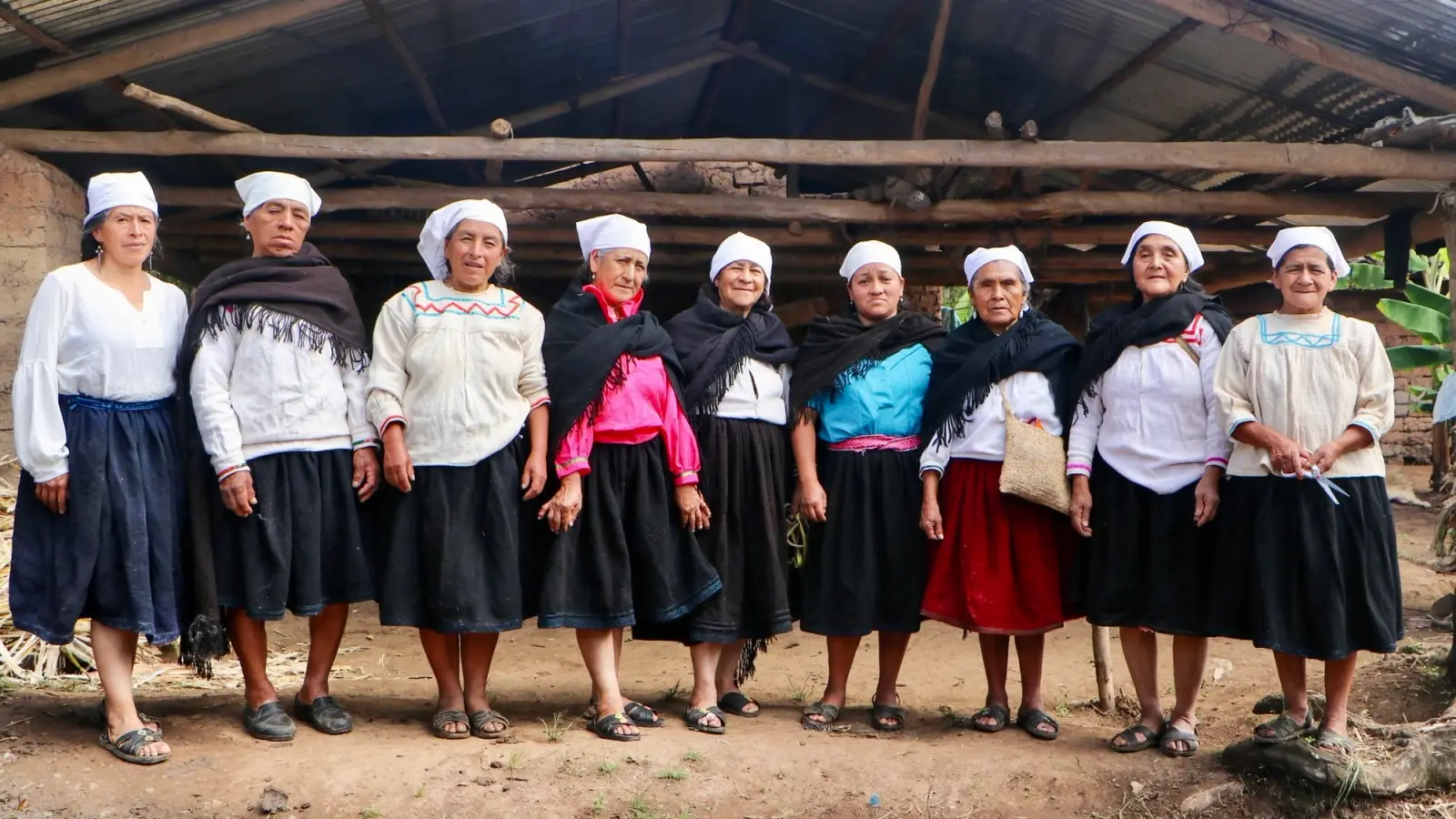 Nueve artesanas con pañuelo blanco y vestimenta tradicional posan bajo un cobertizo